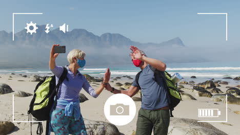 smartphone camera interface screen over caucasian senior couple in face masks on coast taking selfie