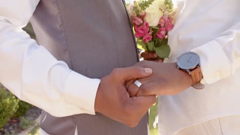 midsection of african american gay male couple holding hands and flowers at wedding, slow motion