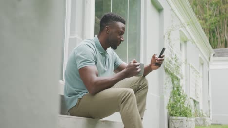happy african american man sitting with smartphone and drinking coffee in garden