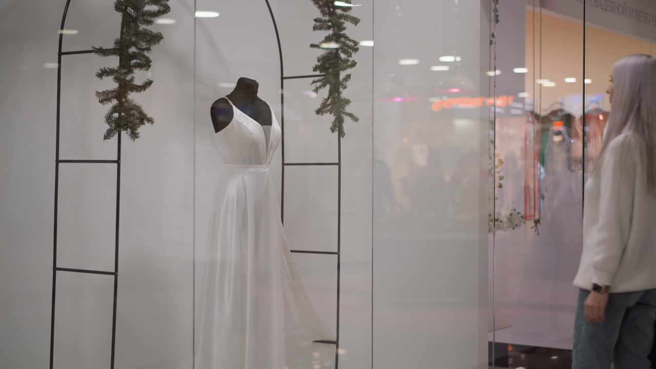 youthful customer carrying bag walks through boutique glass window toward sleeveless long dinner gown on mannequin, elegant silhouette reflected in polished surface under soft store lighting