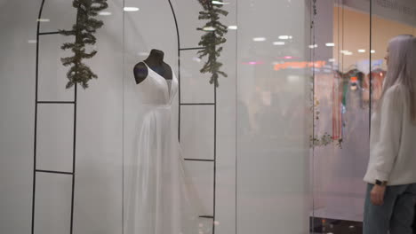 youthful customer carrying bag walks through boutique glass window toward sleeveless long dinner gown on mannequin, elegant silhouette reflected in polished surface under soft store lighting