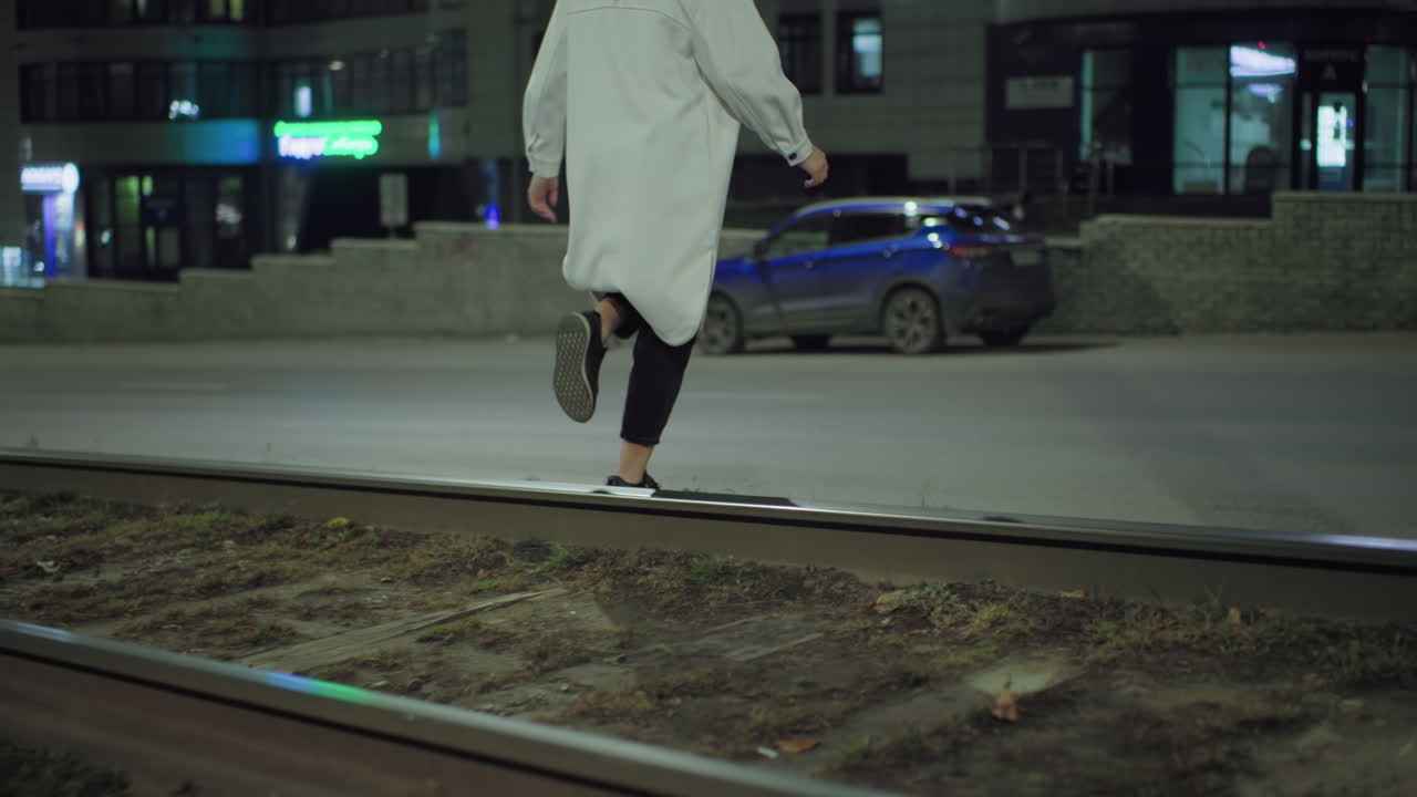 Young woman in long white coat crossing railway track at night toward roadside with parked blue car and residential building in background under glowing city streetlight