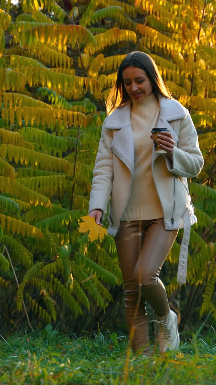 Autumn walk in the beautiful colorful park on warm weather. Gorgeous brunette lady stepping by the grass smiling with a cup in one hand and maple leaf in another.