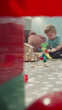 girl holds baby doll while boy playing with robot dinosaur. kids sit on floor near handmade wooden house view through clearance in construction of cubes