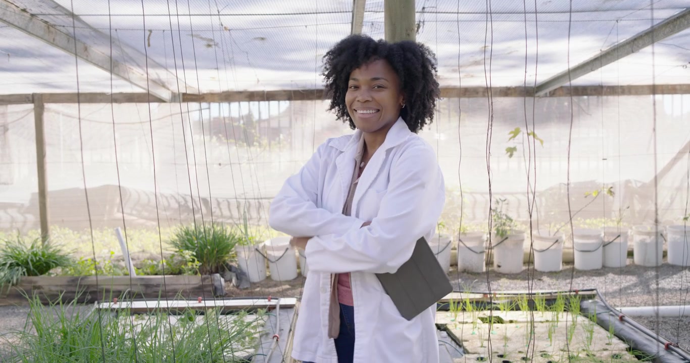 Donna africana che entra in una serra indossando un camice da laboratorio e stivali, usando un tablet per registrare una piantina.
