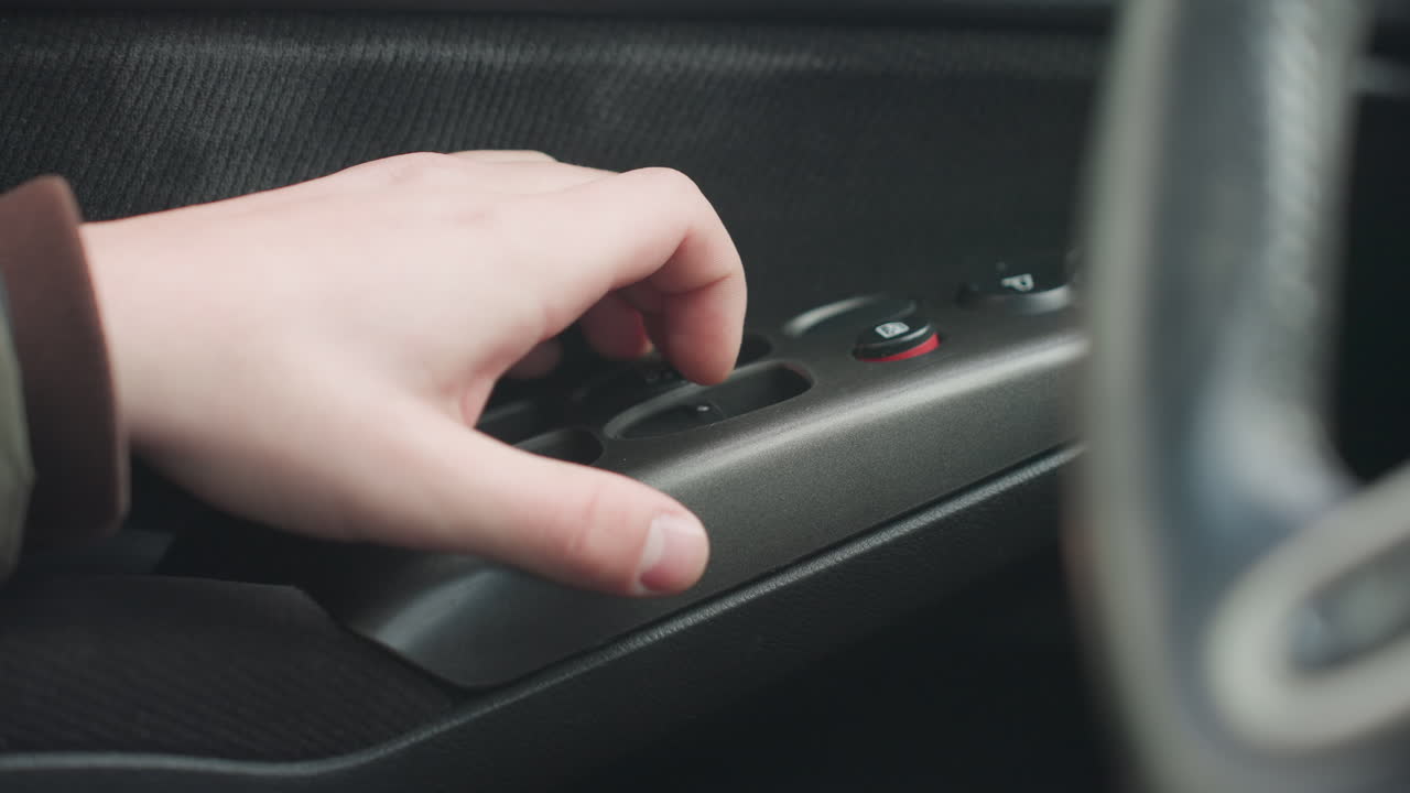 driver hand reaches toward window control buttons on car door panel then pulls away focus on fingers and plastic switches interior lit by daylight showing texture of skin