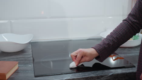 close-up of woman hand wiping glass stove in modern kitchen, brown-covered book rests on countertop while she polishes sleek black surface