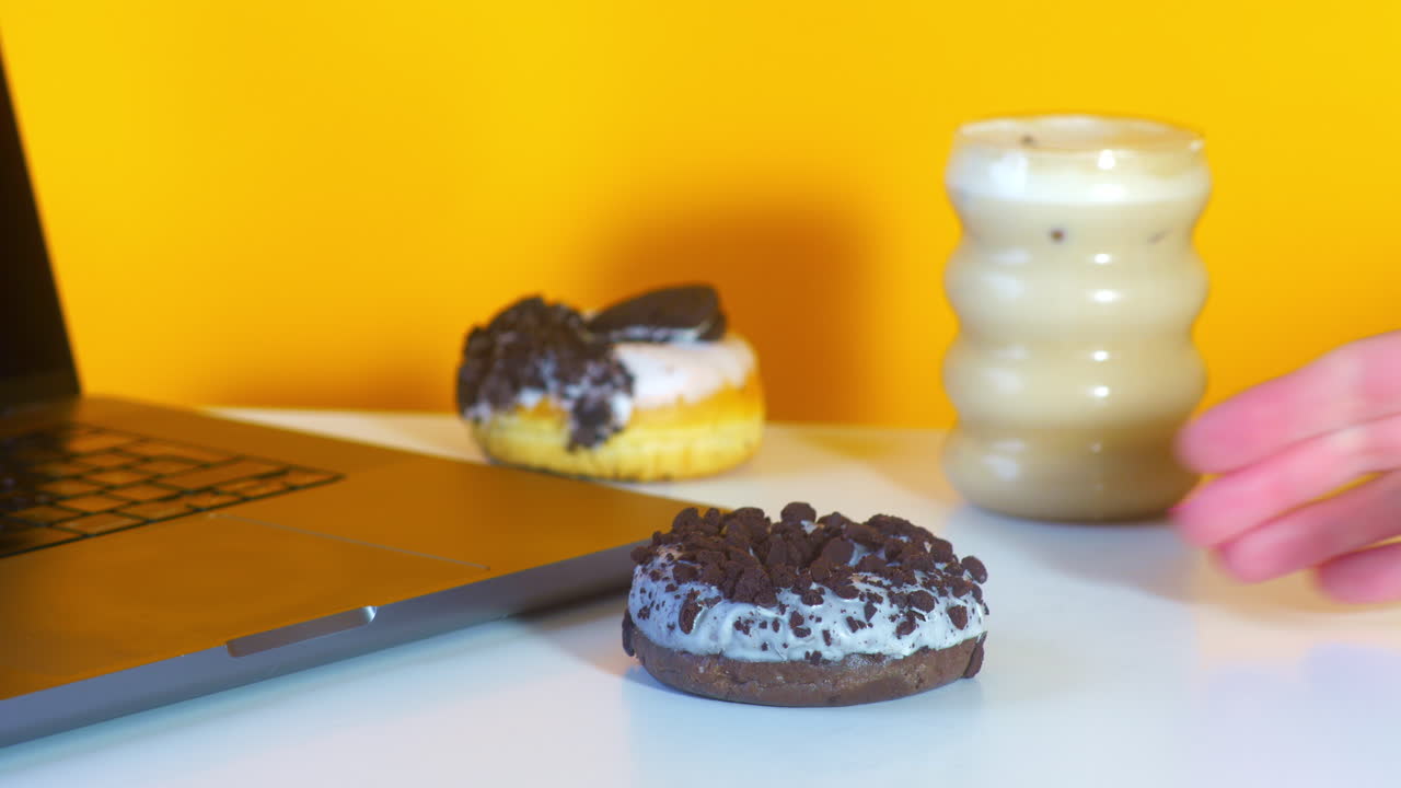 A hand with pink nails grabs a donut topped with cookie crumbles from a desk setup. A laptop, iced coffee, and another donut sit in the background, all set against a bright yellow backdrop.