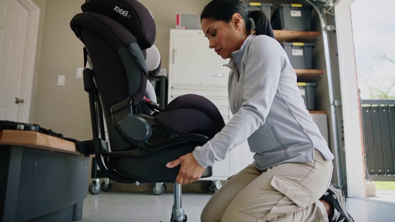 Approaching base woman kneeling aligning securing car seat on wheeled base in garage in zip jacket