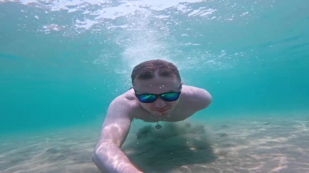 Uomo a torso nudo con occhiali da sole a specchio nuota verso una action cam subacquea, smuovendo sabbia e bolle nel mare greco poco profondo.