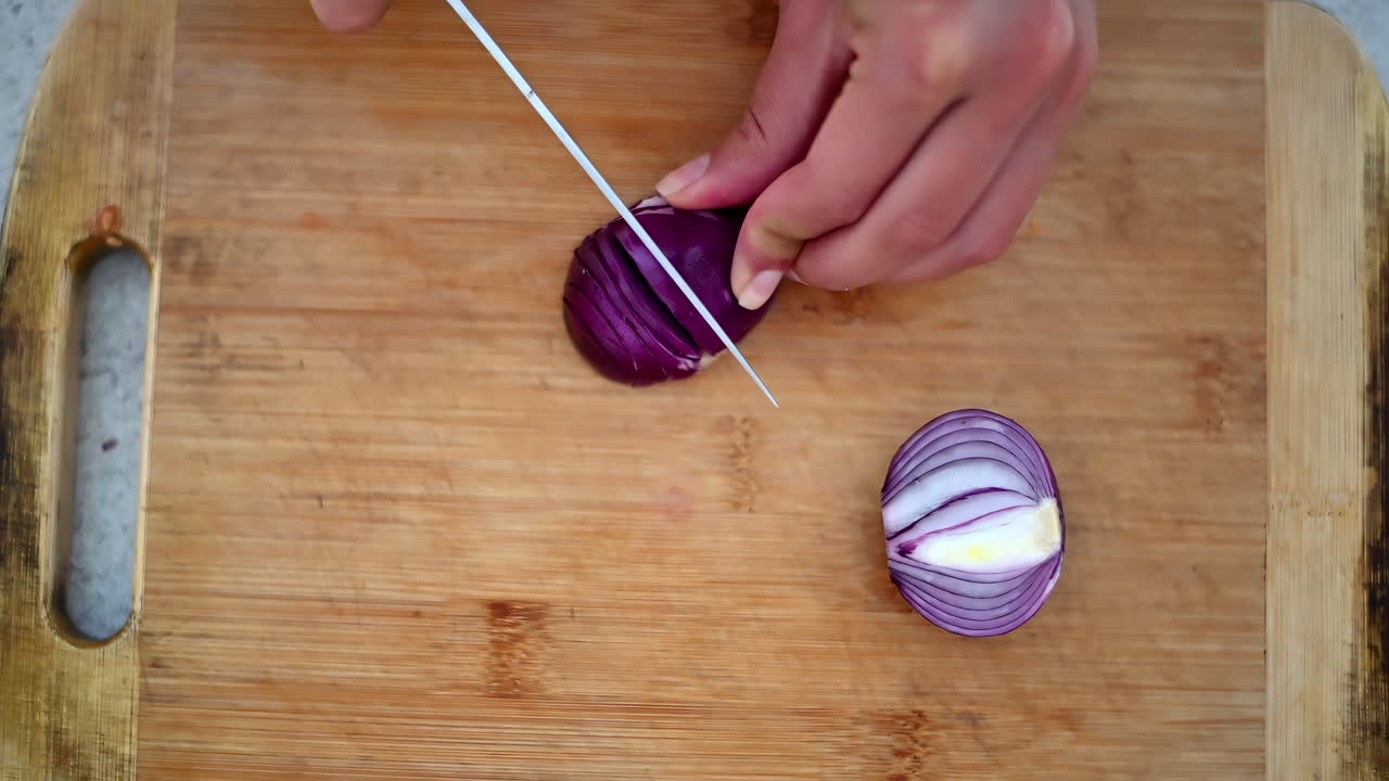 Enjoy seeing a skilled chef expertly slice red onions on a wooden cutting board. The focus is on precise knife skills and preparation in a home kitchen environment