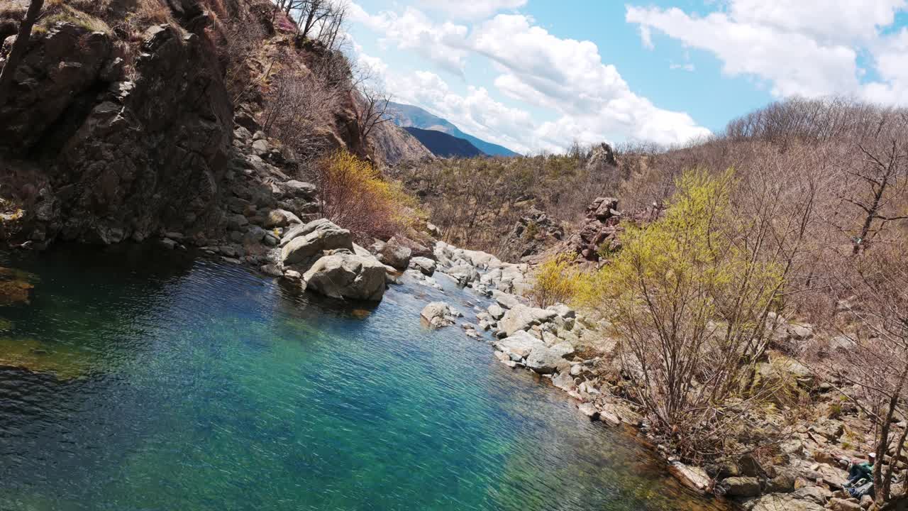 360° rotating shot of a crystal-clear mountain stream flowing through rocky terrain, surrounded by spring foliage and dramatic cliffs under a vibrant blue sky