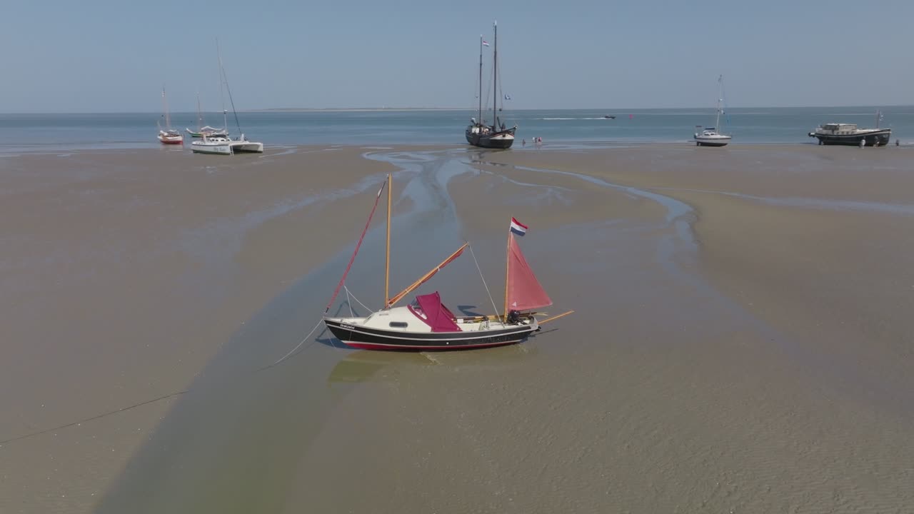 Small red‑sailed dinghy resting on shallow flats with a fleet of boats and a motor barge in the distance; daylight aerial. Wadden Sea