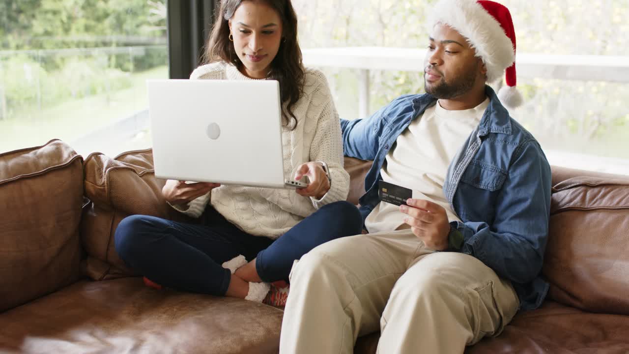 Shifting forward and grabbing laptop, diverse couple on couch completing purchase with credit card