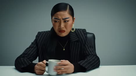 a person sits at a table, holding a mug while displaying a range of emotions. dressed in professional attire, they appear deep in thought in a sleek, minimalist environment.