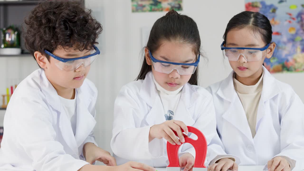 Tiga siswa muda mengenakan kacamata pelindung dan jas lab menjelajahi magnetisme menggunakan magnet tapal kuda bersama
