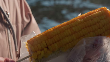 close-up of corn held on stick wrapped in plastic, partially eaten with bokeh light in blurred background, warm steam rising, food photography, snack outdoors