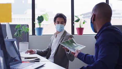 Diverse-businessmen-wearing-face-masks-using-computers-passing-paperwork-in-office