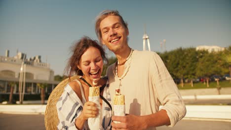 Portrait:-Happy-couple-on-a-date-on-a-modern-beach,-a-blond-guy-with-stubble-in-a-light-shirt-holds-a-hot-dog-along-with-his-girlfriend-who-also-holds-a-hot-dog-and-poses-and-looks-at-the-camera-on-a-date-near-the-sea