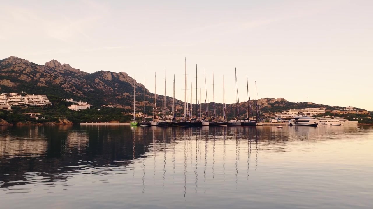 Inquadratura che mostra la marina con yacht ormeggiati e riflessi sull'acqua calma. Porto Cervo in Costa Smeralda, Italia. Inclinazione del drone verso l'alto.