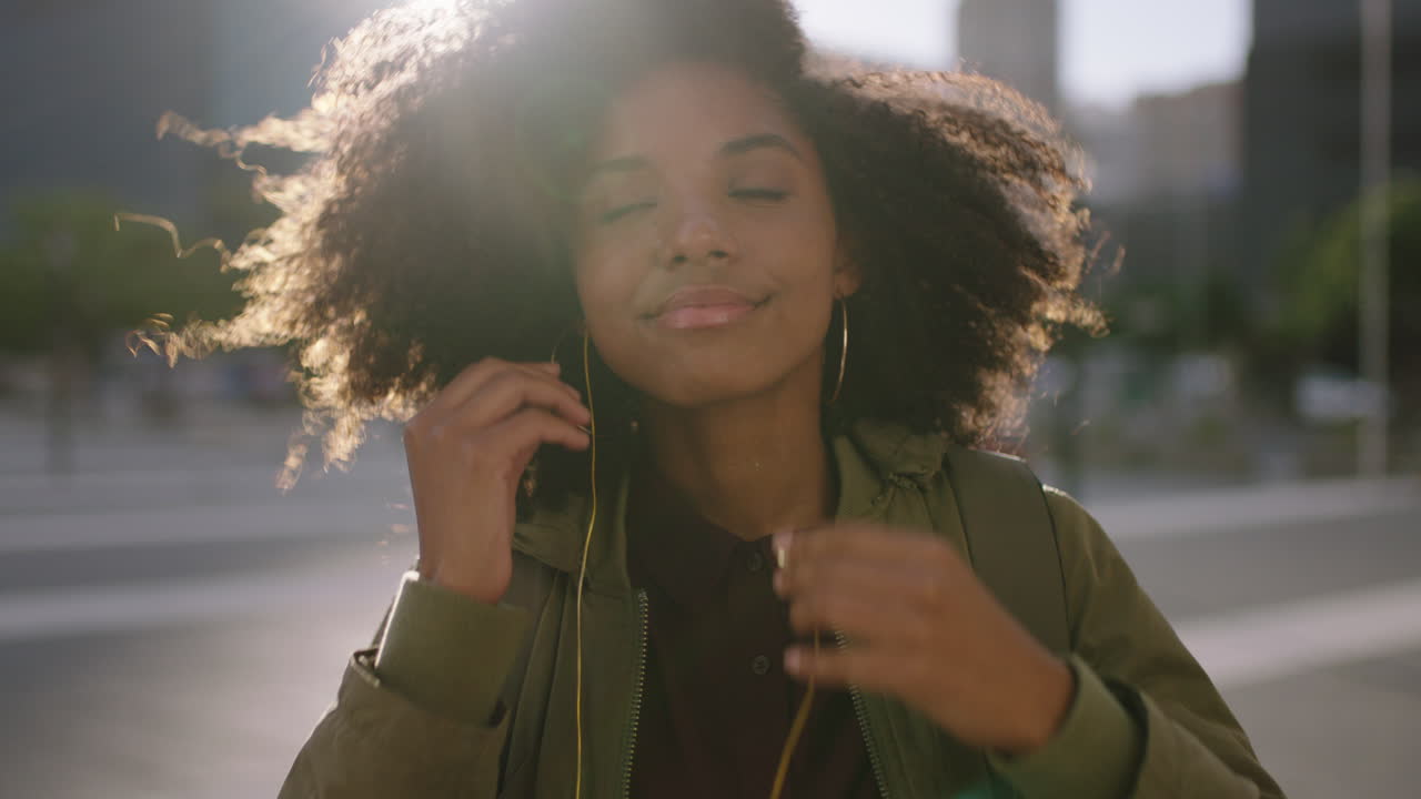 retrato de una hermosa mujer afroamericana de moda se pone auriculares escuchando música disfrutando de la puesta de sol en una ciudad urbana ventosa