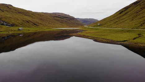 ángulo-Bajo-Aéreo-Con-Alta-Velocidad-Sobre-Un-Hermoso-Lago-Con-Aguas-Tranquilas-Y-Reflectantes-En-Las-Montañas-Junto-A-Una-Carretera-En-Las-Islas-Feroe