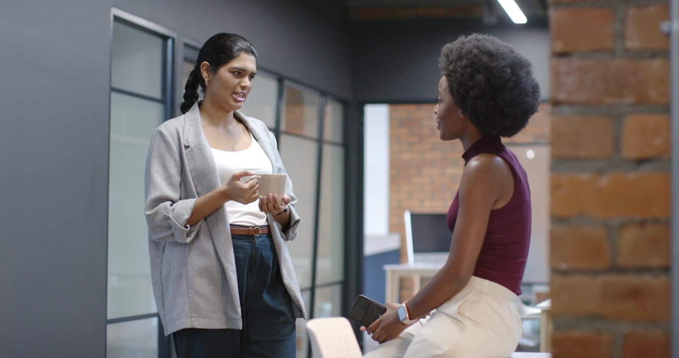 Indian and African American women colleagues holding mug and phone, sharing update in office