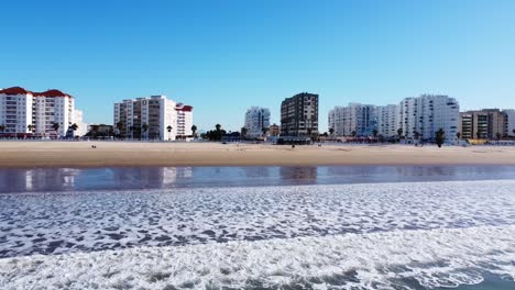 Schöne,-Saubere-Und-Ruhige-Aufnahme-über-Ruhige-Meereswellen-Und-Goldenen-Sandstrand-Mit-Kleinen-Gebäuden