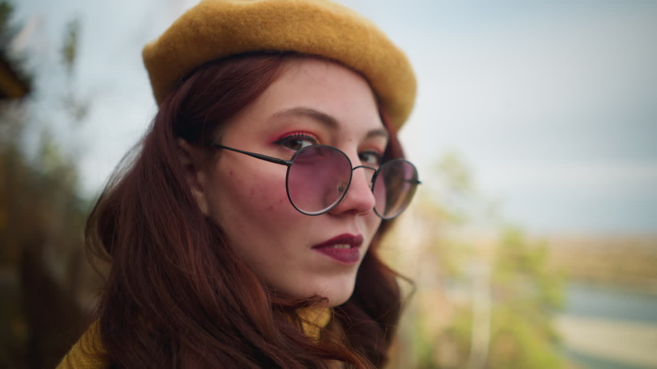 primer plano de una mujer alegre con una boina amarilla y gafas de sol, sus labios pintados curvados en una sonrisa, con un telón de fondo borroso de árboles de otoño, un río y una estructura de madera