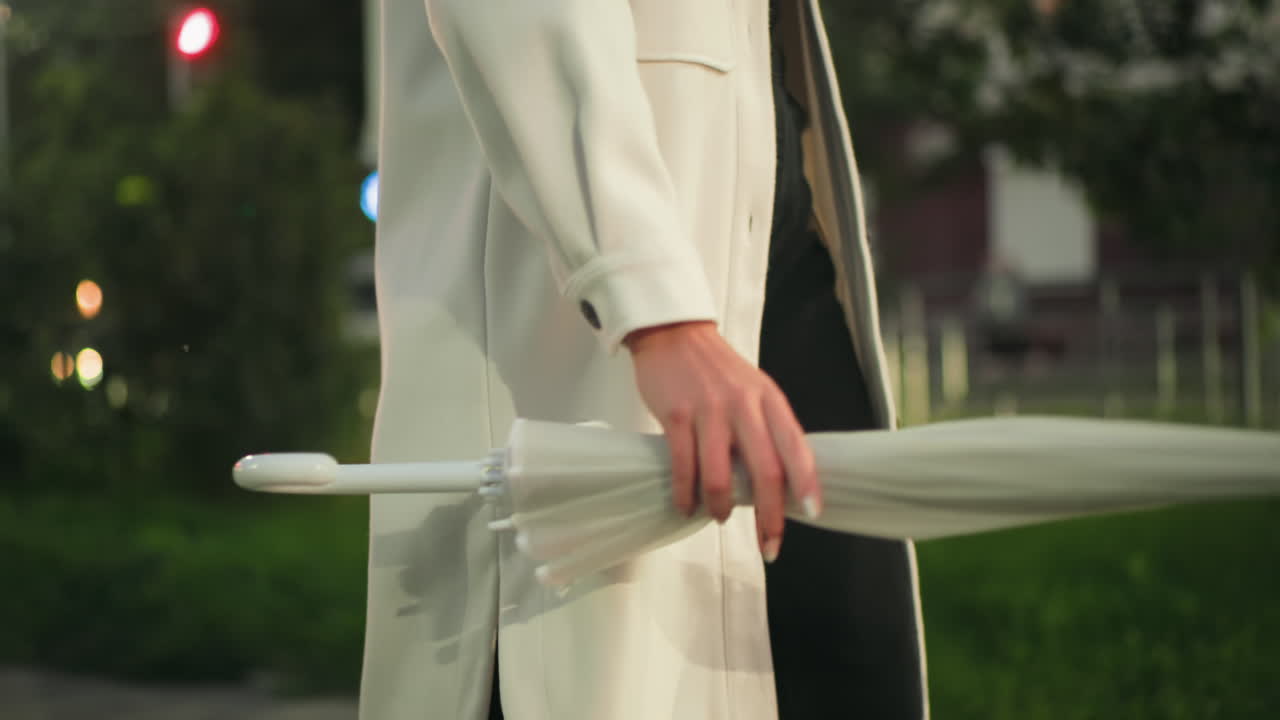 Side partial view of person in white coat swinging folded umbrella during evening stroll with cars passing by, green park background, colorful bokeh lights adding soft glow to urban environment