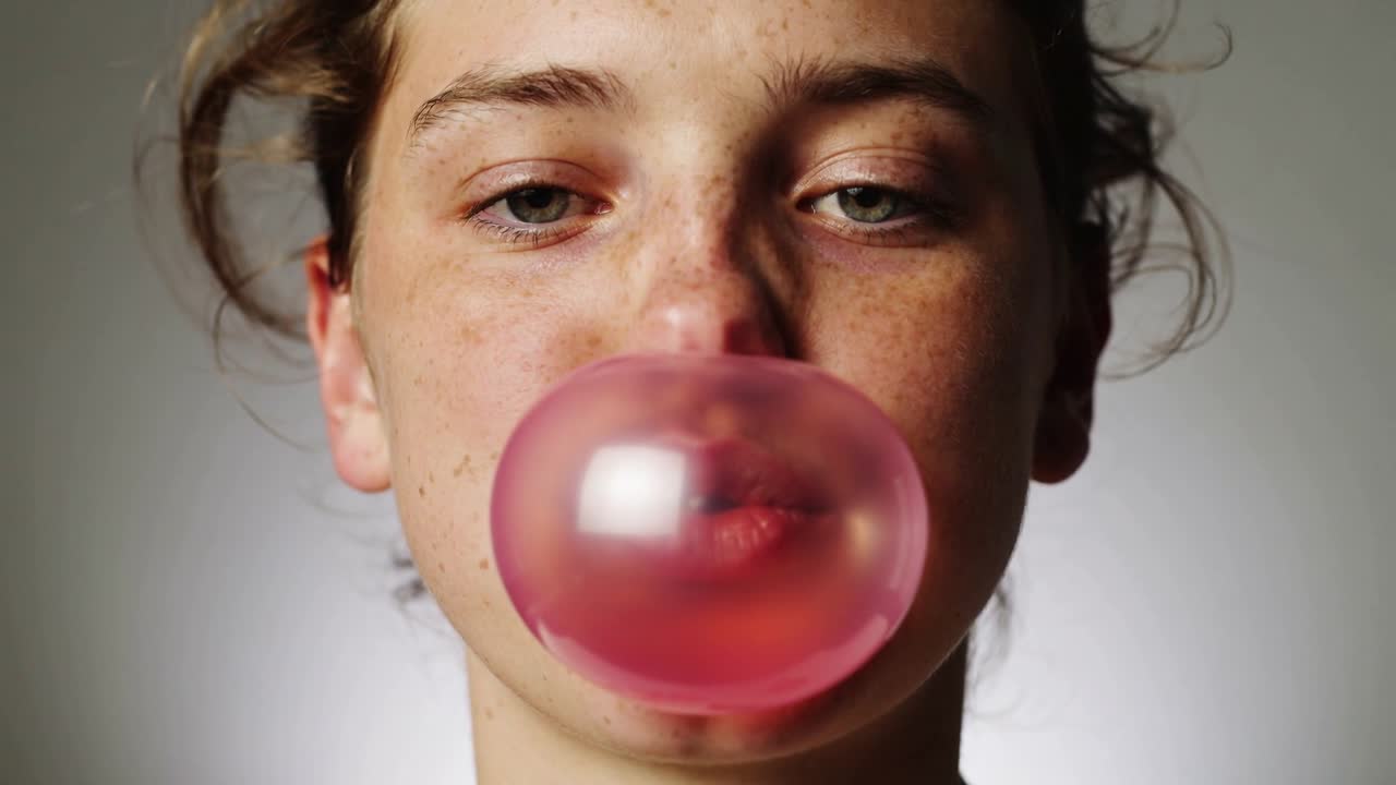 Ein Teenager-Mädchen kaut und bläst dann eine rosa Kaugummiblase vor grauem Hintergrund, die eine große glänzende Kugel bildet.
