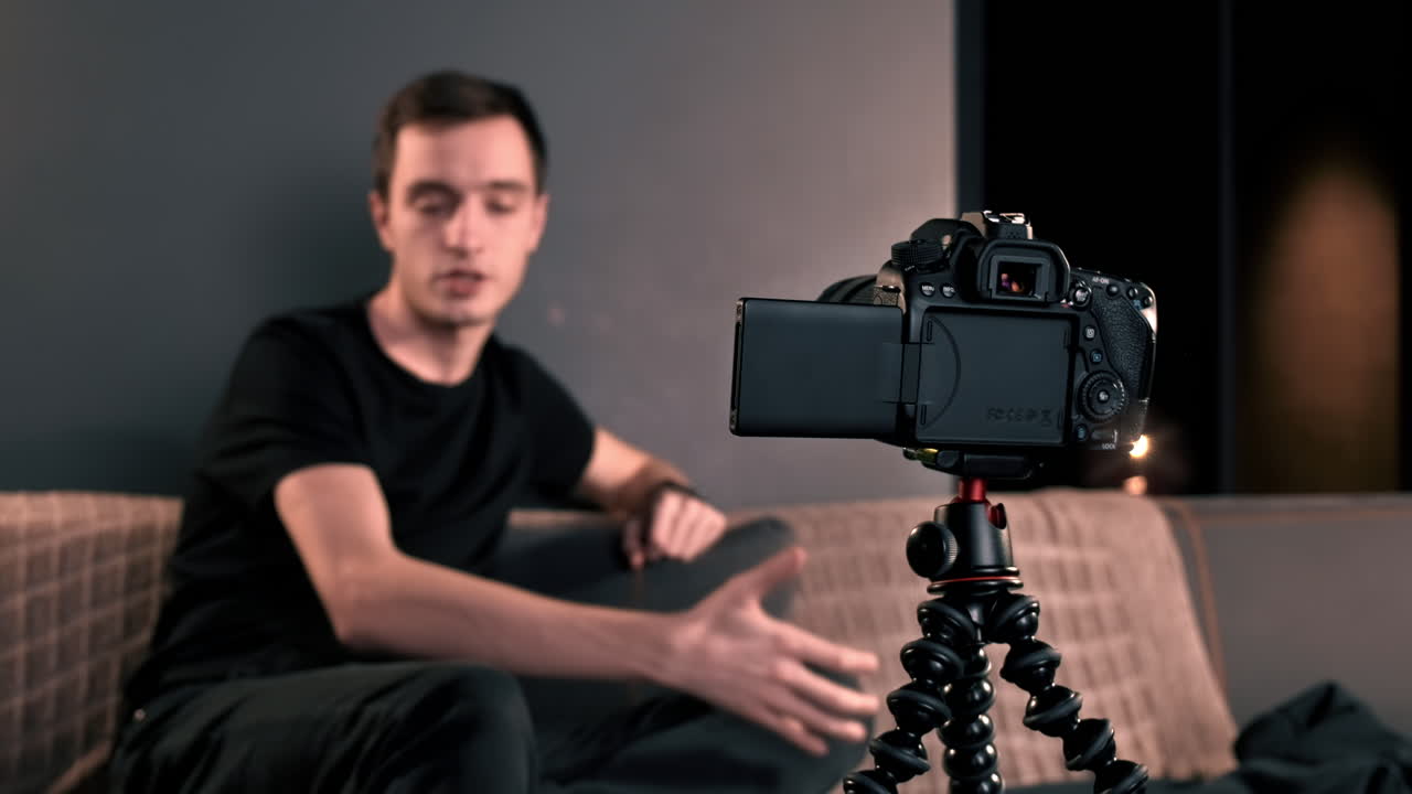 Un giovane creator di contenuti parla e gesticola mentre si riprende usando una fotocamera su un treppiede durante la registrazione di un vlog. Lavora da casa