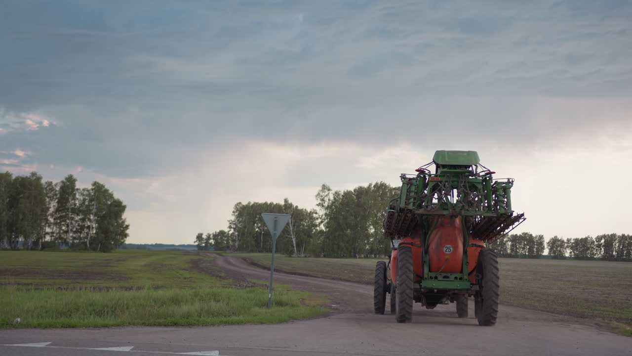 tractor moving along dirt road past post under cloudy sky over green fields beside birch grove, dust trailing from rear wheels, showcasing farming machinery in motion across tranquil rural landscape