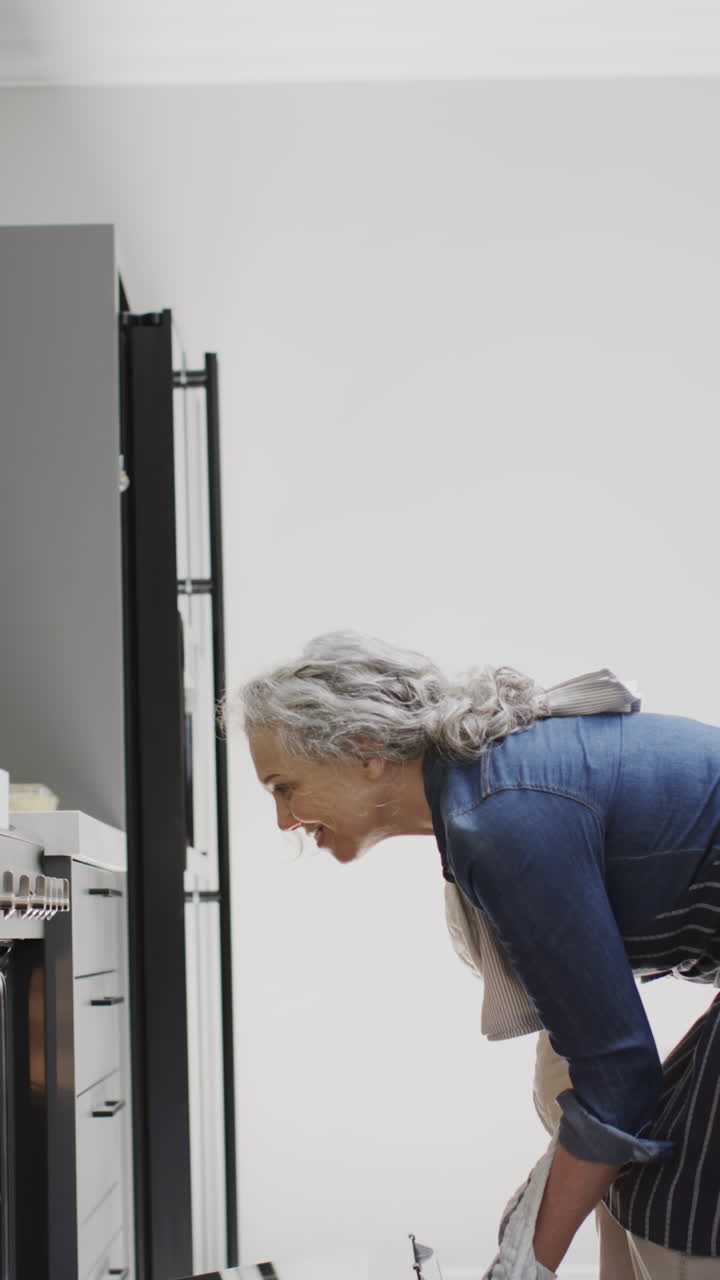Vertical video of senior caucasian woman taking out vegetables from oven in kitchen, slow motion