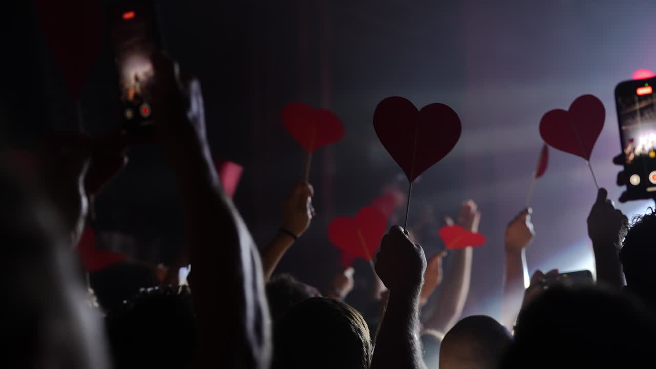 Audience holding red heart shapes during a live concert performance with dramatic lighting effects