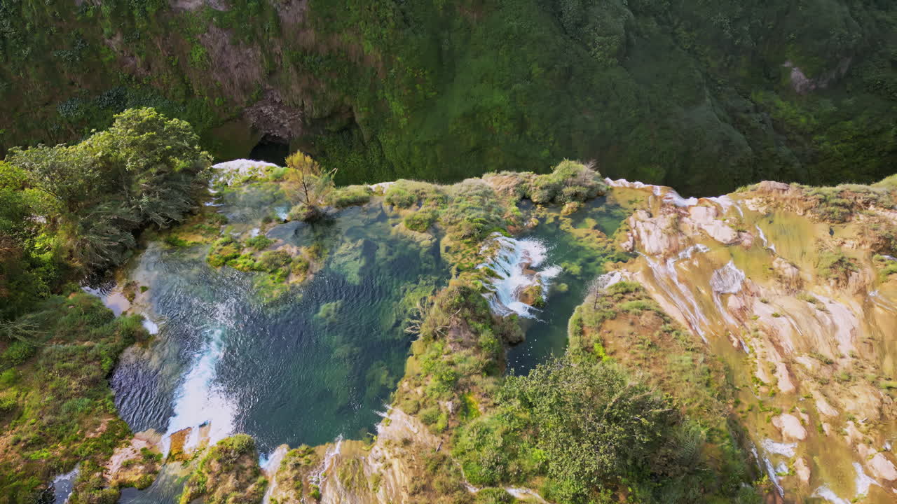 Aerial drone view showing a waterfall flowing into a deep blue basin surrounded by rock formations and vegetation
