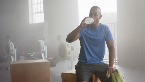 tired biracial man resting and drinking water after training at gym, in slow motion