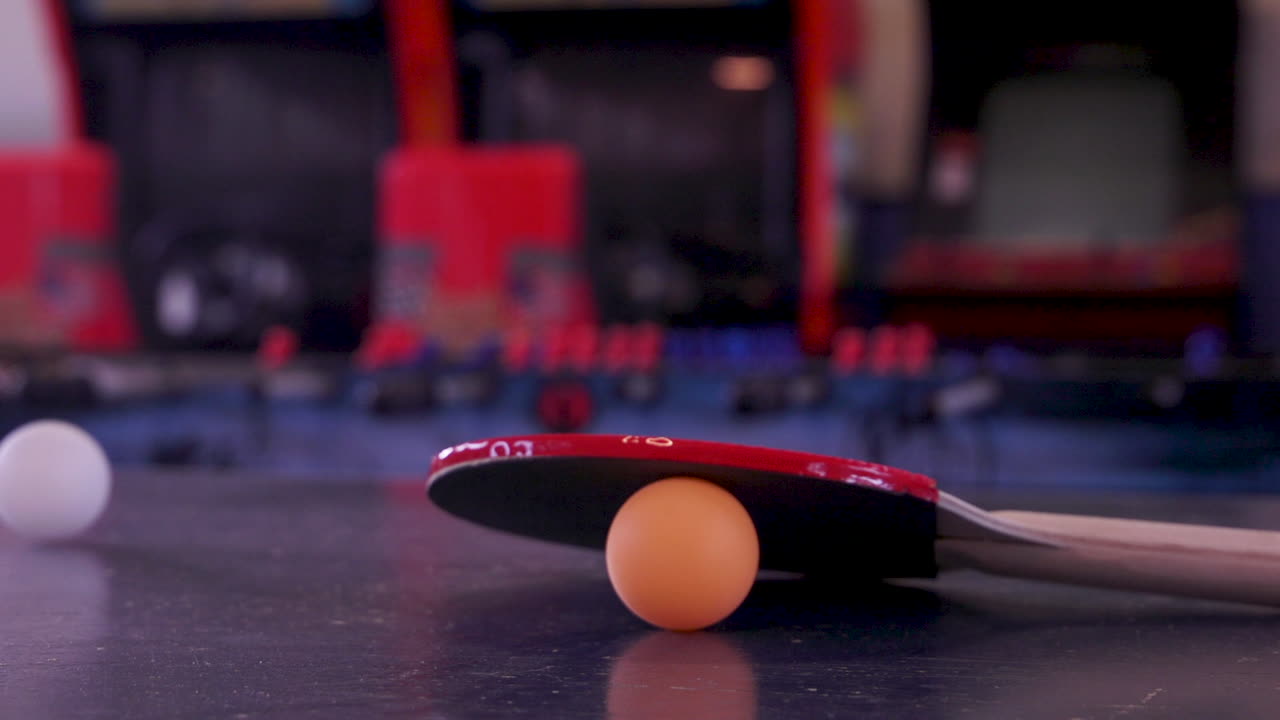 Palla bianca che rotola oltre una racchetta da ping pong rossa e una palla arancione.