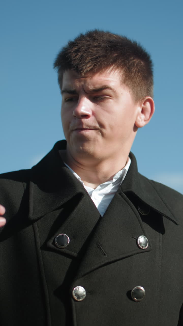 금속 버튼으로 된 검은 코트를 입은 사업가, 밝은 하늘 아래 야외에서 전화 통화를 하고, 흐릿한 현대 건물과 배경의 초록색과 함께 상적으로보고 있습니다.