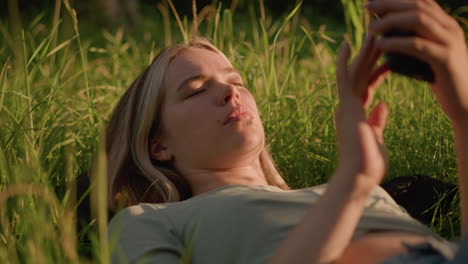 close-up of a woman lying on a grassy field, holding her phone in her hand, with sunlight casting a warm glow on her face and surrounding grass