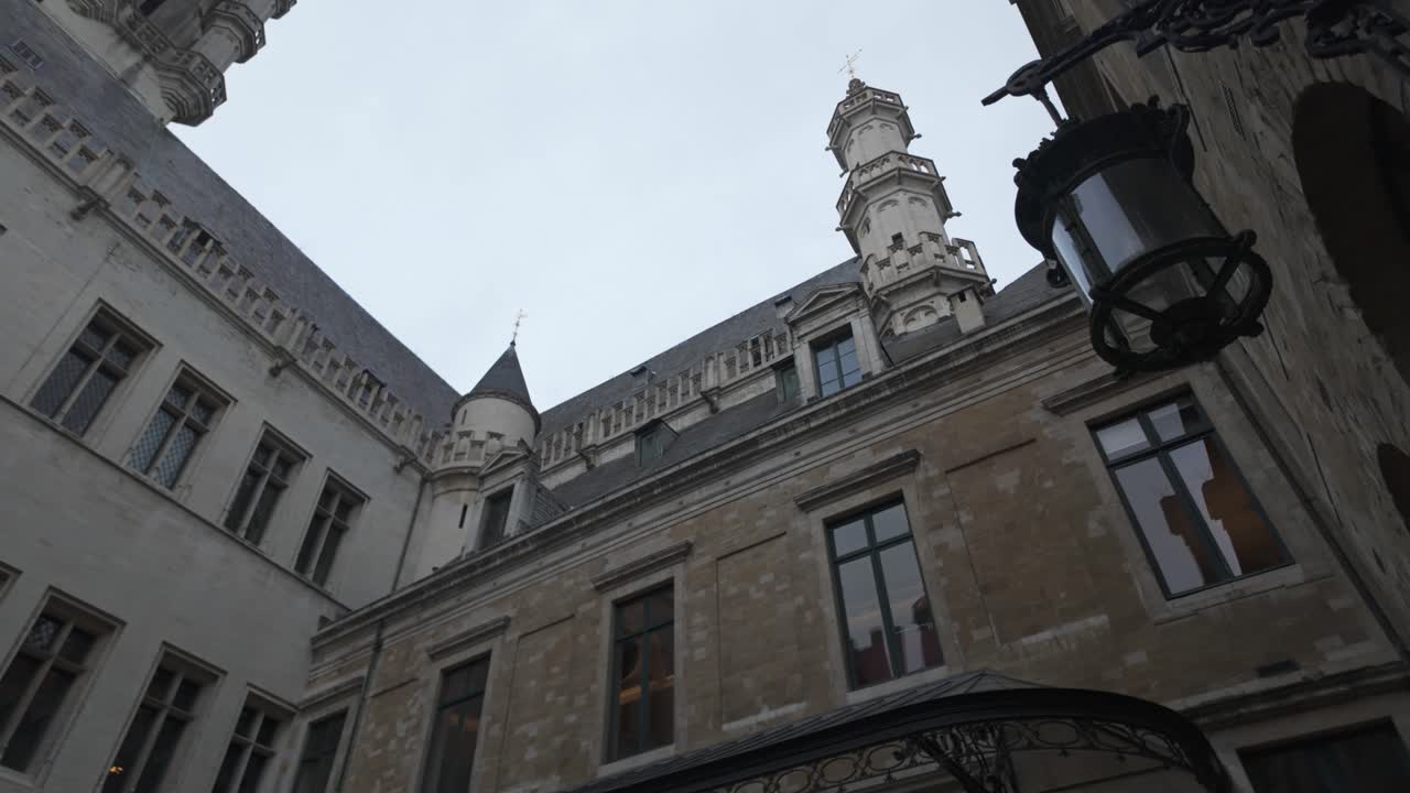 Low angle view of Brussels Town Hall courtyard featuring a hanging lamp, showcasing the gothic architecture and cloudy sky. Pan left