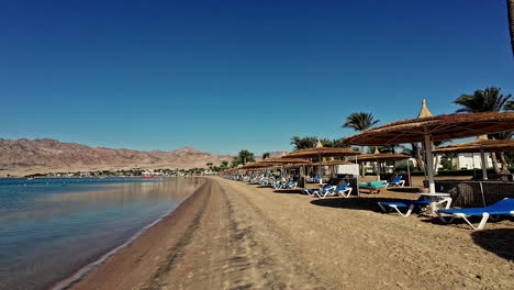 Ruhiger-Strand-Mit-Liegestühlen-Und-Sonnenschirmen,-Klarem-Blauen-Himmel,-Ruhigem-Wasser-Und-Bergkulisse