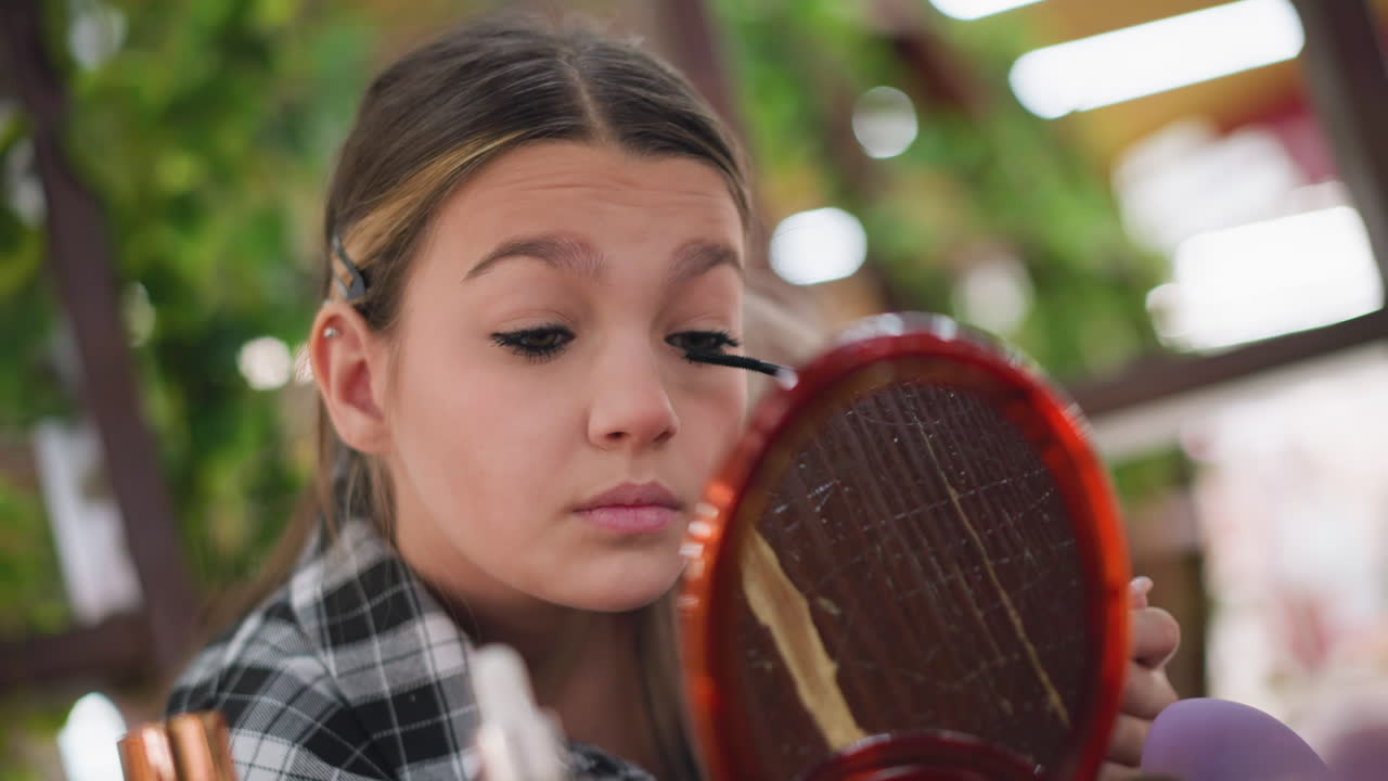 Young lady brushing eyelashes carefully while using a mirror, focusing on eye makeup application, with blurred background of vibrant setting indicating a public space or indoor environment