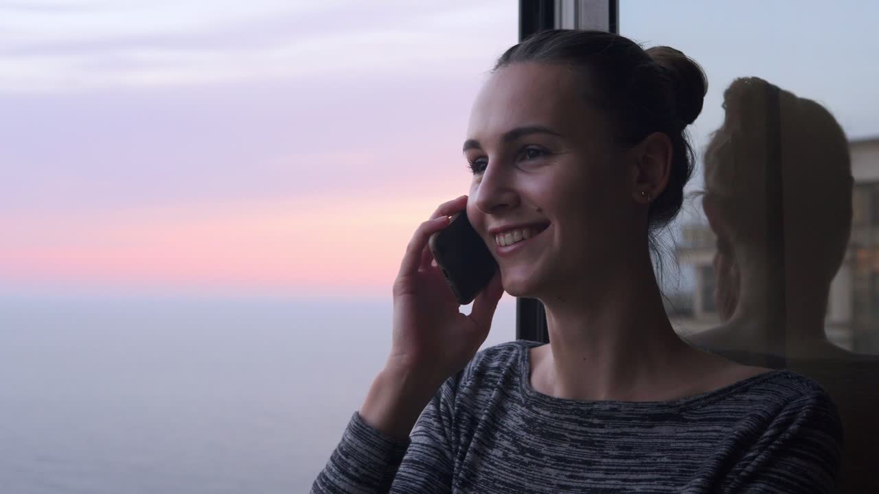 vista en primer plano de una mujer joven sonriente hablando por teléfono mientras estaba de pie junto a la ventana abierta con una sonrisa durante la puesta de sol junto al mar. hermoso cielo y mar en el fondo