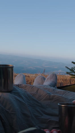 romantic traveler couple lie in tent on mountain hill and drink tea. two tourists rest under blanket during adventure vacation. backpacker family warm up at cold windy weather and admire the scenery.