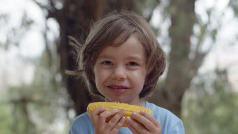 primer plano de niño feliz comiendo mazorcas de maíz y mirando a la cámara