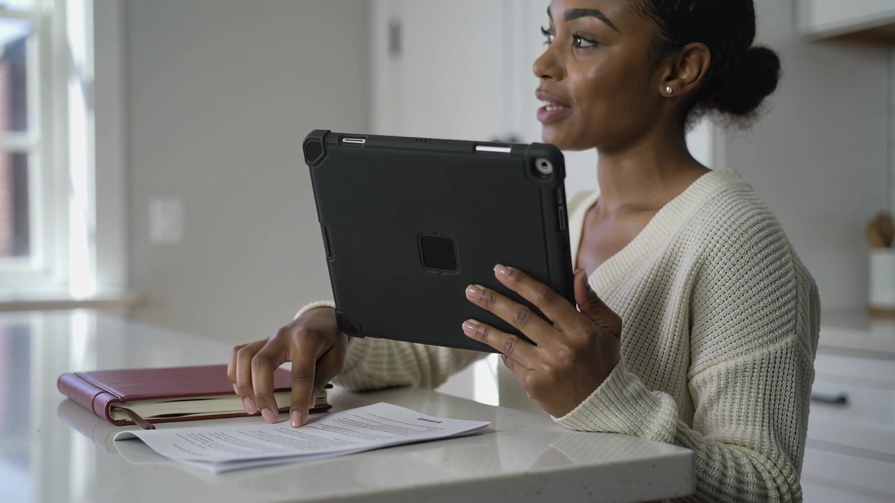 Holding tablet, woman in knit sweater joining call in kitchen, reviewing papers, maroon notebook