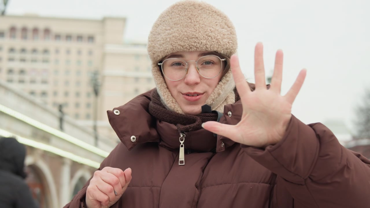 Young woman stands on city promenade in winter, adjusting jacket cuffs and gloves with animated hand gestures while smiling toward camera, wearing knit hat, glasses, and brown puffer jacket beside