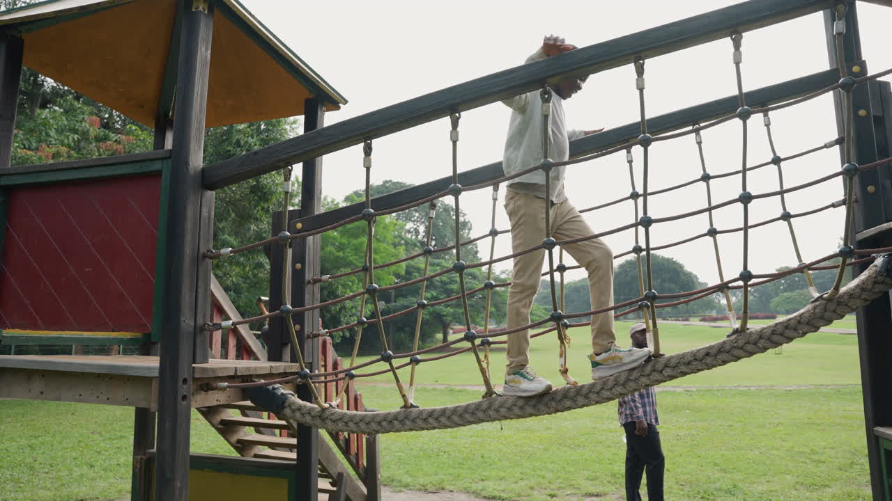Father crossing rope bridge watched by son, carefully balancing on wooden walkway above open grass, child stands below looking up, protective teaching moment with teamwork and guidance in park, candid