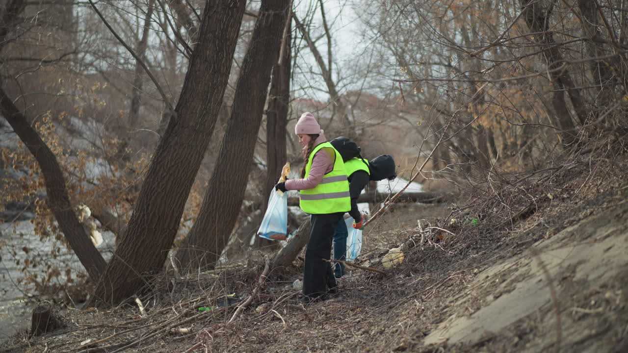 Two volunteers wearing reflective safety vests and gloves picking up trash and debris from riverbank, placing waste into blue bags during outdoor cleanup to protect environment, reduce pollution